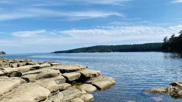 Smooth flat rocks line the edge of a calm blue lake, with forested hills in the distance under a bright sky filled with scattered clouds. Peaceful scene.
