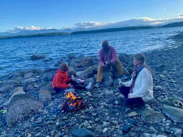 Three people sit around a campfire on a rocky beach, enjoying drinks. The blue water, distant islands, and mountains create a peaceful, scenic backdrop.