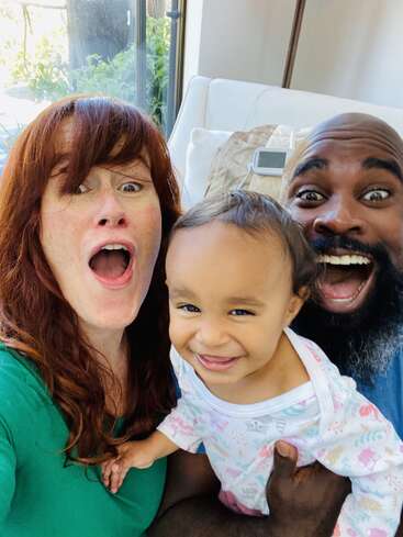 A joyful family selfie featuring a red-haired woman, bearded man, and a smiling baby. Their happy expressions radiate warmth, love, and fun in a cozy home.