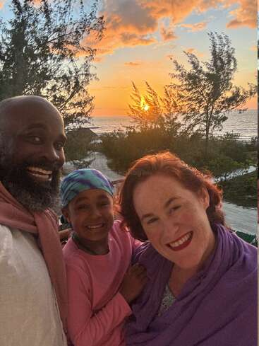 Une famille de trois personnes, tout sourire, profite d'un magnifique coucher de soleil sur la plage, avec des arbres, des sentiers de sable et l'océan en arrière-plan, immortalisant ainsi un moment de vacances joyeux passés ensemble.