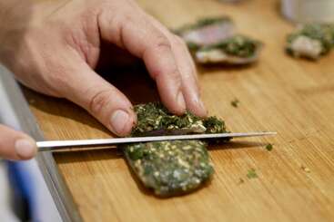 Eine Hand schneidet mit einem scharfen Messer ein Stück Fleisch oder Fisch mit Kräuterkruste auf einem hölzernen Schneidebrett. Andere Stücke sind in der Nähe zu sehen.