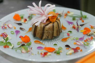 Un plat gastronomique magnifiquement présenté avec un élément central moulé, garni de tourbillons artistiques de sauces, de légumes éclatants, de fleurs comestibles et de microgreens disposés de manière décorative.