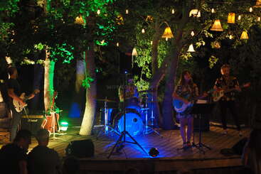 Un groupe de musique se produit sur une scène extérieure en bois éclairée par des lumières colorées, entourée d'arbres et de lampes suspendues, sous le regard d'un public au premier plan, la nuit.