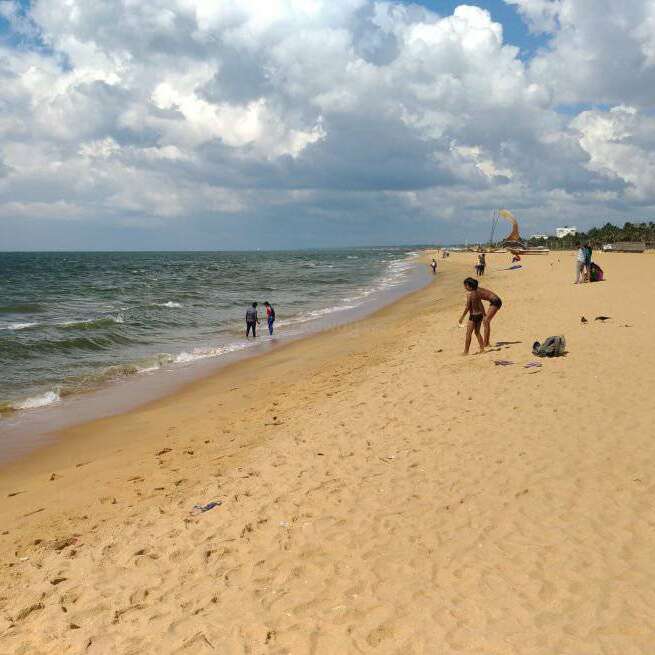 Das Bild zeigt eine ruhige Strandszene mit einem Sandstrand, sanften Wellen und ein paar Menschen, die unter einem bewölkten Himmel spazieren gehen oder sich entspannen.