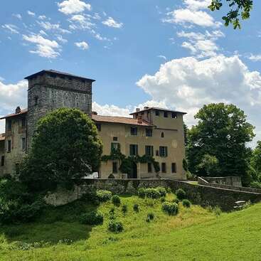 L'image représente un grand bâtiment à plusieurs étages avec une tour en pierre, entouré d'une végétation luxuriante et d'un mur en pierre, sur fond de ciel bleu avec des nuages blancs.