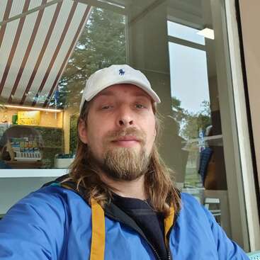 L'image montre un homme aux cheveux longs et à la barbe, portant une casquette blanche et une veste bleue, prenant un selfie devant une fenêtre et un magasin.