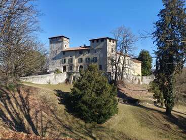 L'image représente un grand bâtiment en pierre à plusieurs étages avec une tour, situé sur une colline entourée d'arbres et d'un ciel bleu clair.