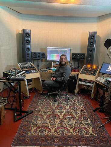 L'image représente un homme assis dans un studio d'enregistrement, entouré de divers équipements et instruments de musique, avec un tapis sur le sol et un écran d'ordinateur derrière lui.