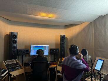 L'image représente deux personnes dans un studio d'enregistrement, entourées de matériel de musique, l'une portant des écouteurs et l'autre se tenant à un microphone.