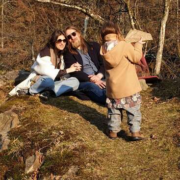 A imagem mostra uma família de três pessoas sentadas na grama em uma área arborizada, com uma criança em pé na frente deles, segurando uma pedra.