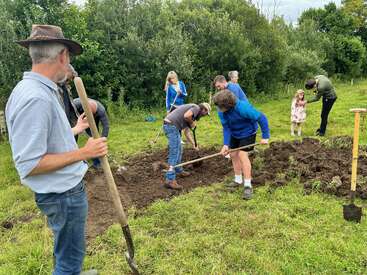 Un grupo de personas, entre las que hay adultos y niños, están al aire libre cavando y trabajando con palas en un terreno cubierto de hierba, rodeado de árboles y vegetación verde.