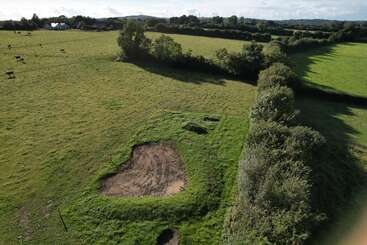 Vista aérea de un campo rural verde con vacas pastando, atravesado por setos. En primer plano, una excavación con tierra expuesta y manchas de barro.