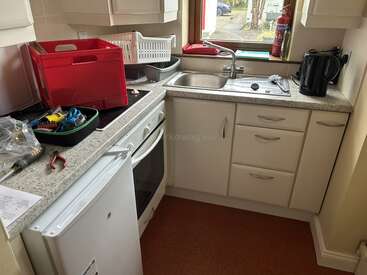 This compact kitchen features white cabinets, a small oven, mini fridge, sink, dish rack, kettle, and countertop tools, with a window providing natural light.