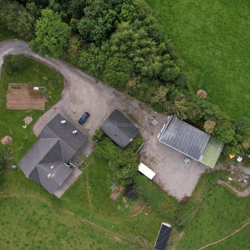 Dieses Luftbild zeigt ein ländliches Grundstück mit drei Gebäuden, einer Einfahrt, Bäumen, Grasflächen, geparkten Fahrzeugen, verstreuten Außengeräten und einer umgebenden Straße, von oben gesehen.