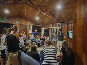 A group of young people gathers in a cozy wooden room, some seated, others standing, engaging in conversation or listening attentively to two presenters.