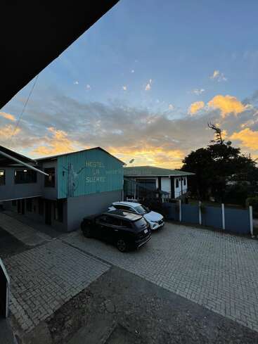 A peaceful hostel parking lot at sunrise or sunset, with three cars parked. The sky is colorful with bluish hues and scattered orange clouds overhead.