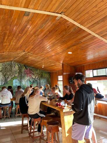 A group of people are gathered around wooden tables in a cozy room with a wooden ceiling, enjoying food and conversation. A mural decorates the back wall.
