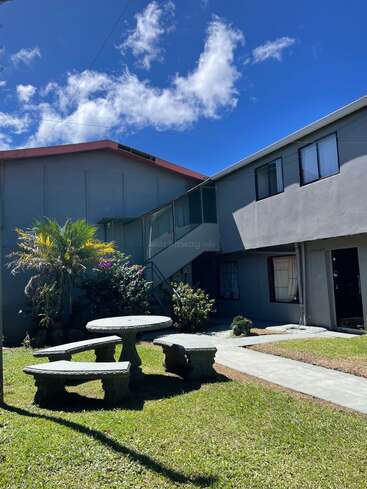 This image shows a sunny courtyard with green grass, a stone picnic table and benches, plants, bushes, and two-story gray buildings under a blue sky.