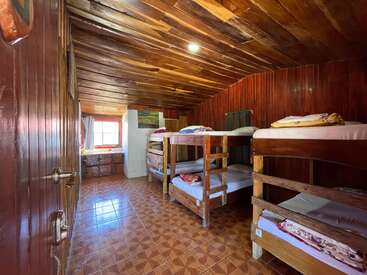 This image shows a cozy, rustic dormitory room with four wooden bunk beds, tiled floor, wooden paneled walls and ceiling, and sunlight streaming through a window.