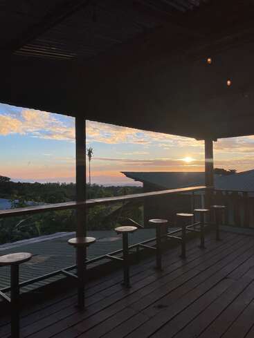 A peaceful sunset view from a wooden balcony with empty bar stools, overlooking rooftops and lush greenery under a colorful sky filled with soft clouds.
