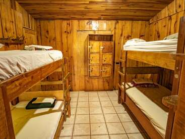 This cozy wooden room features two sets of bunk beds, tiled floors, warm lighting, and a small storage cabinet, creating a rustic and inviting cabin atmosphere.