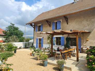 A charming house with blue shutters, rustic beige walls, and a tiled roof. The patio features a wooden pergola, outdoor furniture, and potted plants in sunlight.