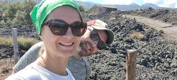 A smiling couple takes a selfie outdoors. The woman wears sunglasses and a green headscarf. Behind them, volcanic rocks, a rustic building, and distant mountains are visible.