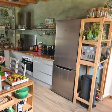 This cozy kitchen features wooden shelves, modern appliances, a stainless steel fridge, plants, organized jars, and a table with fresh fruits and vegetables, creating a warm atmosphere.