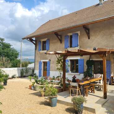 Una casa con encanto, con contraventanas azules, paredes rústicas de color beige y un tejado de tejas. El patio cuenta con una pérgola de madera, muebles de exterior y plantas en macetas bañadas por la luz del sol.