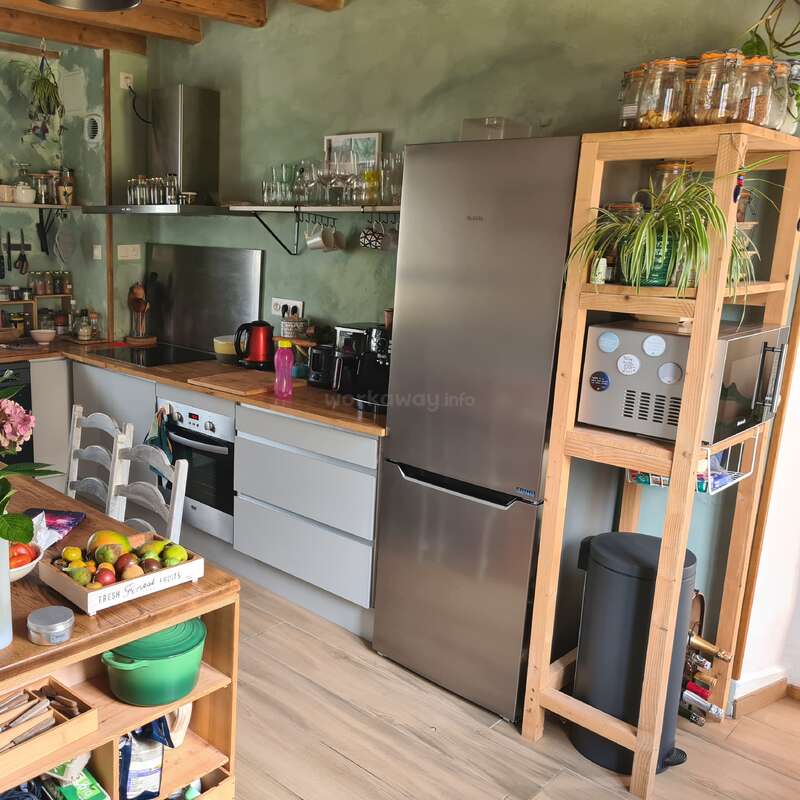 This cozy kitchen features wooden shelves, modern appliances, a stainless steel fridge, plants, organized jars, and a table with fresh fruits and vegetables, creating a warm atmosphere.
