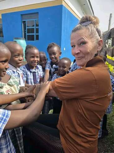 A group of smiling school children in uniform gather around a woman in a brown shirt, outside a blue building, enjoying a joyful and playful moment together.