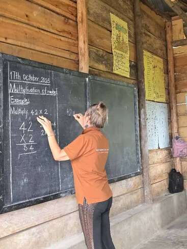 A teacher writes multiplication examples on a blackboard in a rustic classroom with wooden walls, educational posters, and backpacks hanging. The atmosphere feels focused and educational.