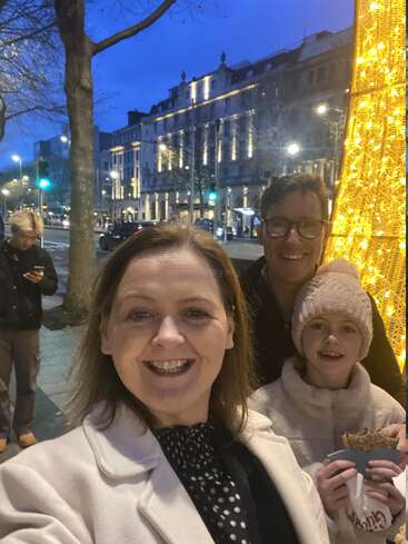 Une famille souriante prend un selfie près de lumières festives dans une rue de la ville au crépuscule. L'enfant tient de la nourriture et les bâtiments brillent chaleureusement en arrière-plan.