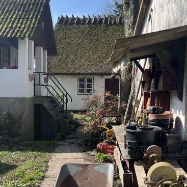 The image depicts a serene rural scene featuring a white house with a moss-covered roof, surrounded by lush greenery and a stone pathway leading to a charming garden area.