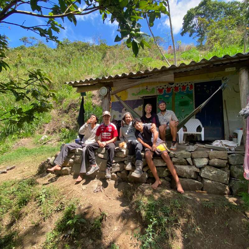 Cinq personnes sont assises sur un muret devant une maison rustique, entourées de verdure, d'arbres et d'un soleil radieux. Elles semblent détendues et heureuses.
