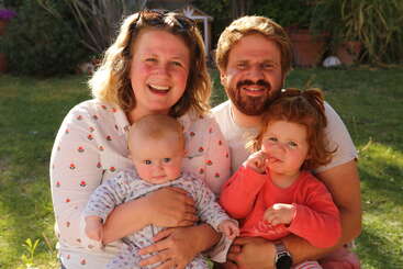 Eine glückliche vierköpfige Familie sitzt im Freien im Sonnenlicht. Die Mutter hält ein Baby im Arm, während der Vater ein Kleinkind umarmt, und alle lächeln sich herzlich an.