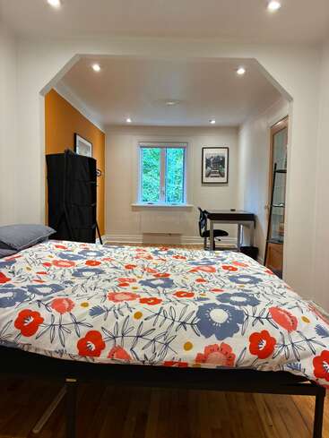 Ein gemütliches Schlafzimmer mit geblümter Bettwäsche, einem Holzboden, einem Schreibtisch am Fenster, heller Beleuchtung, einer orangefarbenen Akzentwand, einem Kleiderschrank und gerahmter Wandkunst.
