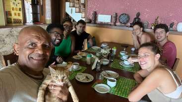 Eine Gruppe von sieben Leuten sitzt um einen Esstisch herum und lächelt für ein Selfie. Der Mann vorne hält eine orangefarbene Katze im Arm. Es herrscht eine fröhliche, ausgelassene Stimmung.