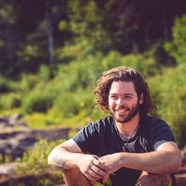 A imagem mostra um homem com longos cabelos castanhos, vestindo uma camiseta azul-escura, sentado ao ar livre em um ambiente natural com vegetação e árvores ao fundo.