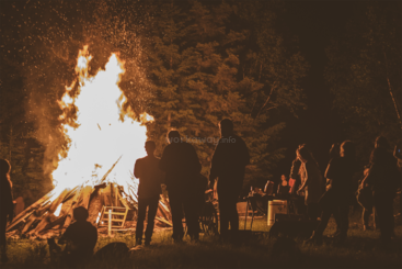 L'image représente un groupe de personnes réunies autour d'un grand feu, probablement à l'occasion d'un événement social, avec un chemin dégagé et une clôture à l'arrière-plan, ce qui donne un sentiment de communauté et de rassemblement.