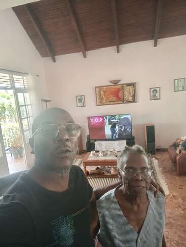 A man and an elderly woman pose together in a cozy living room, with wooden ceiling, TV, framed pictures, coffee table, and outdoor light coming through windows.