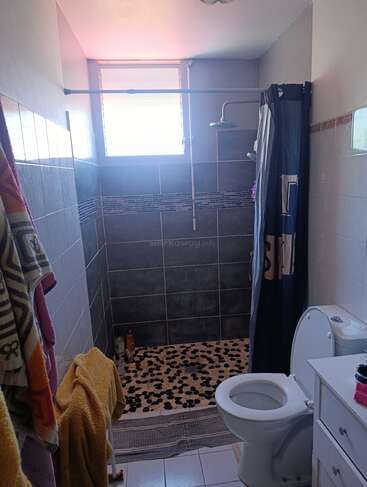 This is a bathroom with a walk-in shower featuring pebble flooring, grey tiles, a window, toilet, white vanity, and colorful towels hanging on the wall.