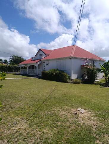 A charming white house with a red roof sits under a partly cloudy sky, surrounded by green grass, shrubs, and tropical plants, exuding tranquility.