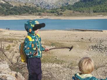 Deux enfants explorent les rives rocheuses d'un lac. L'un d'eux, vêtu d'un sweat à capuche coloré et d'un sac à dos jaune, tient des bâtons. L'eau bleue et les montagnes créent un décor paisible.