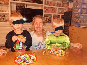 Eine lächelnde Frau sitzt zwischen zwei kleinen Jungs mit Snacks. Die Jungs haben ihre Augen bedeckt; einer trägt ein Superheldenkostüm. Um sie herum liegen bunte Teller und Spielsachen.