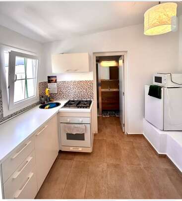 Il s'agit d'une petite cuisine moderne équipée d'armoires blanches, d'un dosseret en mosaïque, d'une cuisinière à gaz, d'une fenêtre ouverte, d'un réfrigérateur, d'un micro-ondes et d'un sol en carrelage brun clair, qui mène à une salle de bains.