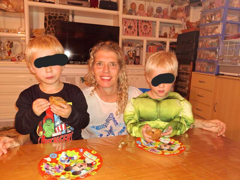 Une femme souriante est assise entre deux jeunes garçons avec des collations. Les garçons ont les yeux bandés ; l'un d'eux porte un costume de super-héros. Des assiettes colorées et des jouets les entourent.