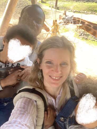 A family takes a cheerful selfie at a zoo, with giraffes visible in the background. The children’s faces are blurred for privacy, creating a joyful moment.