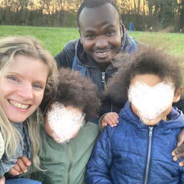 Uma família sorridente de quatro pessoas está sentada ao ar livre, na grama verde. Os pais estão bem próximos um do outro, enquanto os rostos das crianças estão desfocados por motivos de privacidade. Há árvores ao fundo.