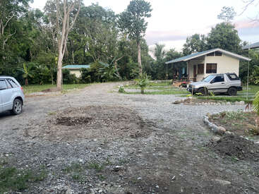 A small house with a porch and parked car is surrounded by lush greenery, gravel driveway, trees, and another visible vehicle in a peaceful rural setting.